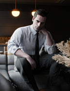 Genesis 9 Male character seated on a sofa in a dimly lit lounge setting, wearing a light gray micro-check dress shirt with a dark tie and charcoal pinstripe trousers, hand resting thoughtfully against his face.